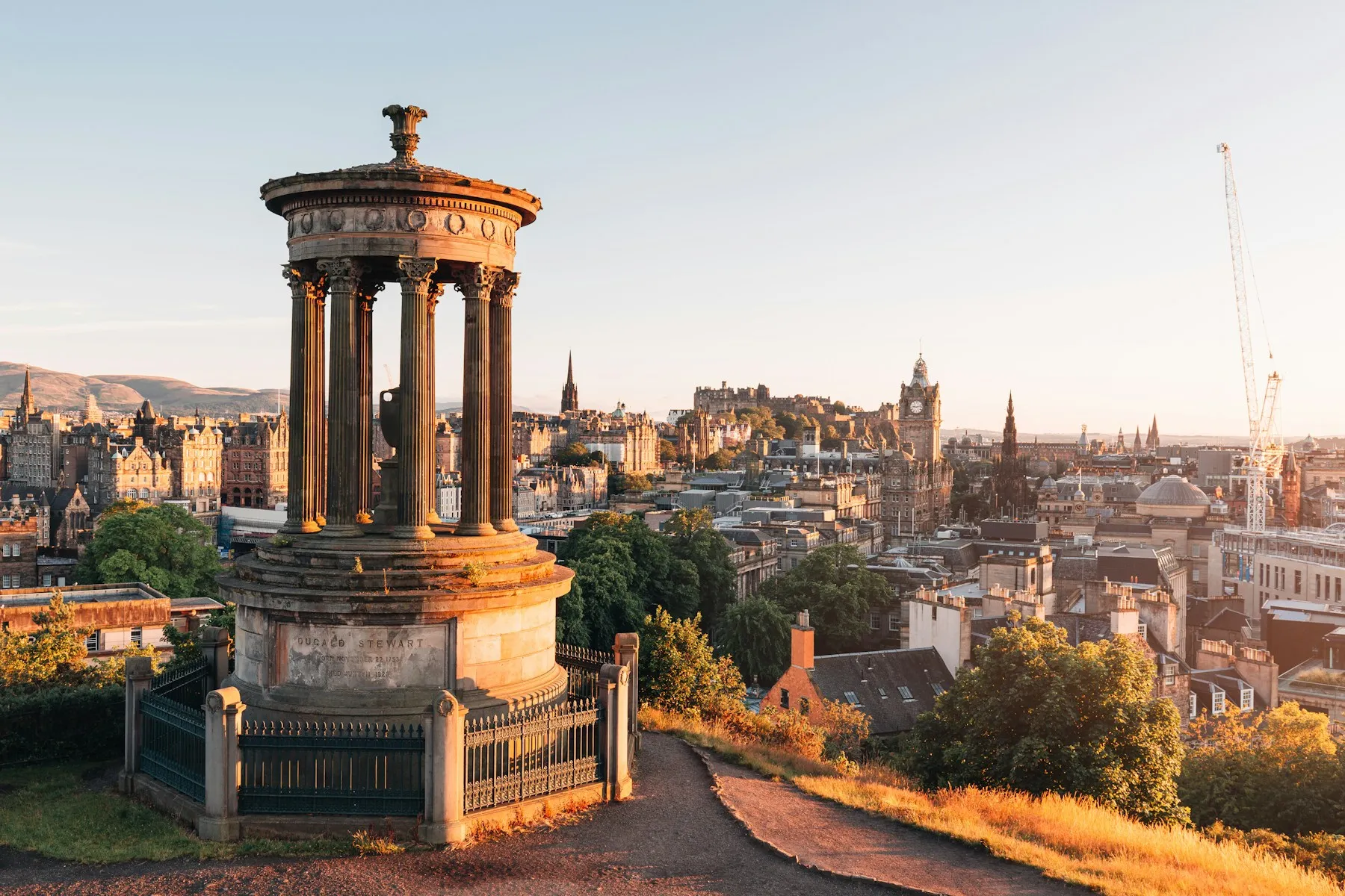 The circular colonnade of the Dugald Stewart Monument on Calton Hill at sunset, with Edinburgh Castle and the city skyline beyond.