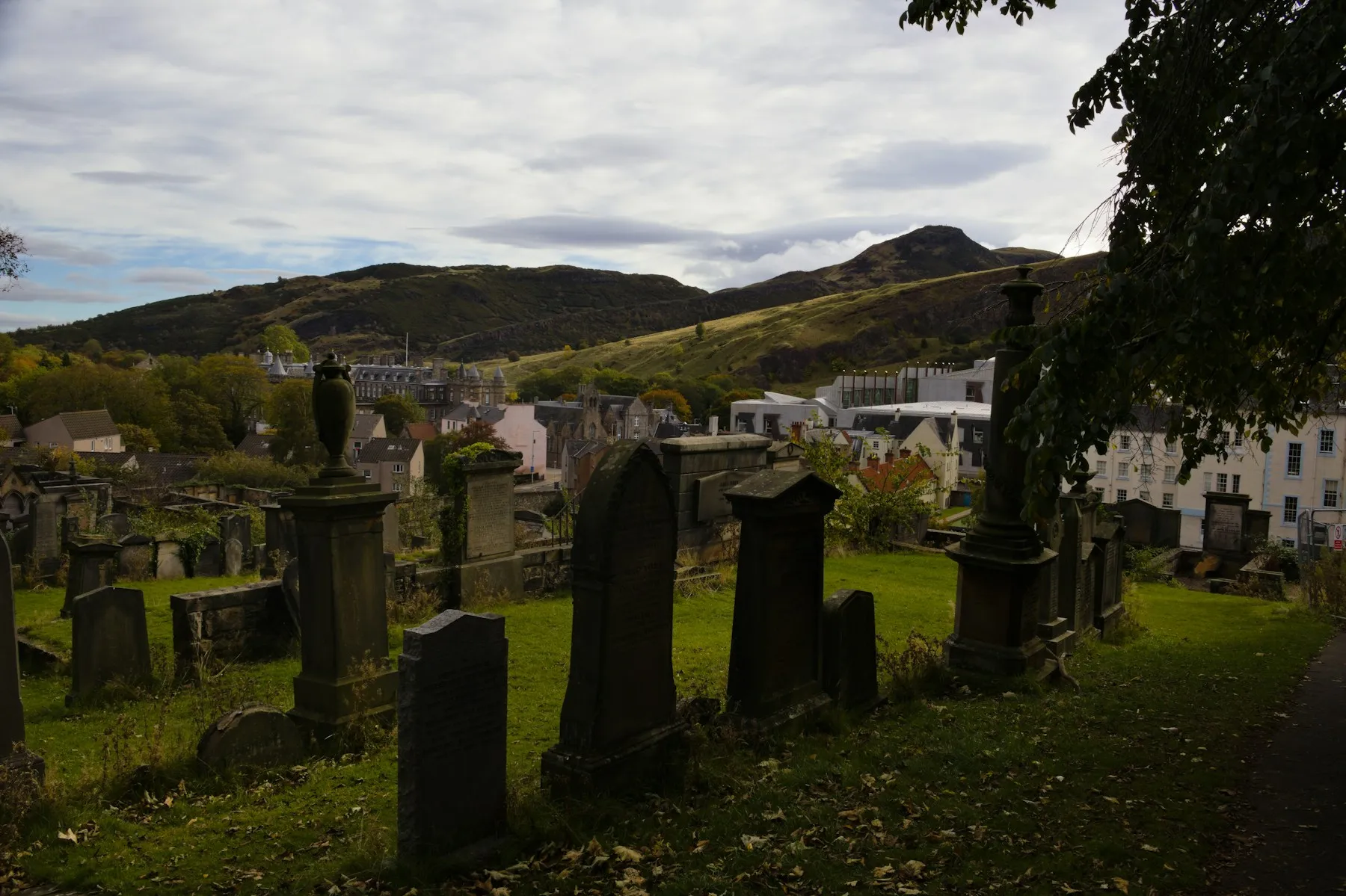 Weathered gravestones and stone monuments in a historic Edinburgh burial ground, with Arthur's Seat rising behind the rooftops.