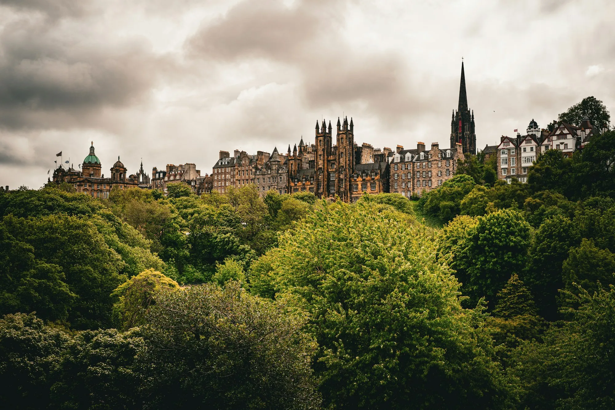Edinburgh's Old Town skyline of spires and historic stone buildings rising above dense trees under a heavy grey sky.