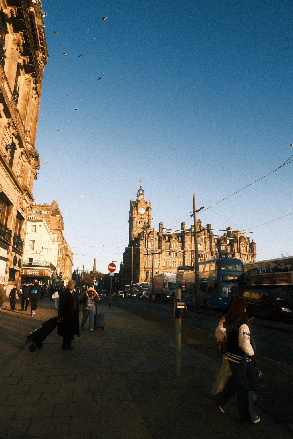 Princes Street in Edinburgh in low golden light, with the Balmoral clock tower at the far end and birds scattering above the rooftops.