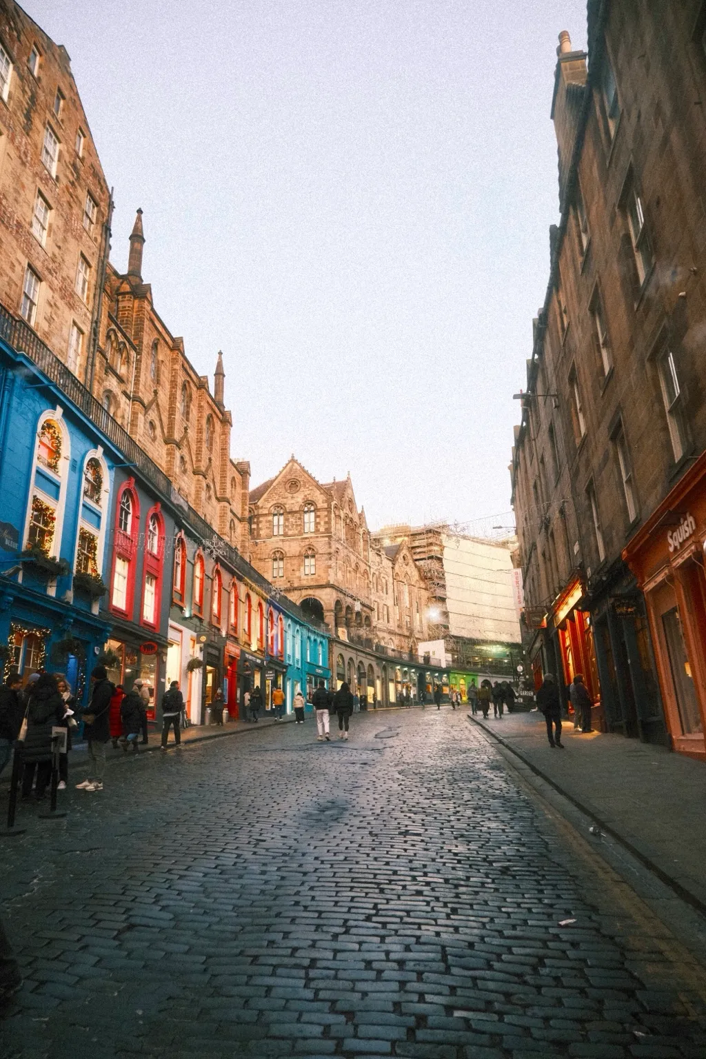 The curved cobbled slope of Victoria Street in Edinburgh, lined with blue, red and green shopfronts, wet setts reflecting the winter light.